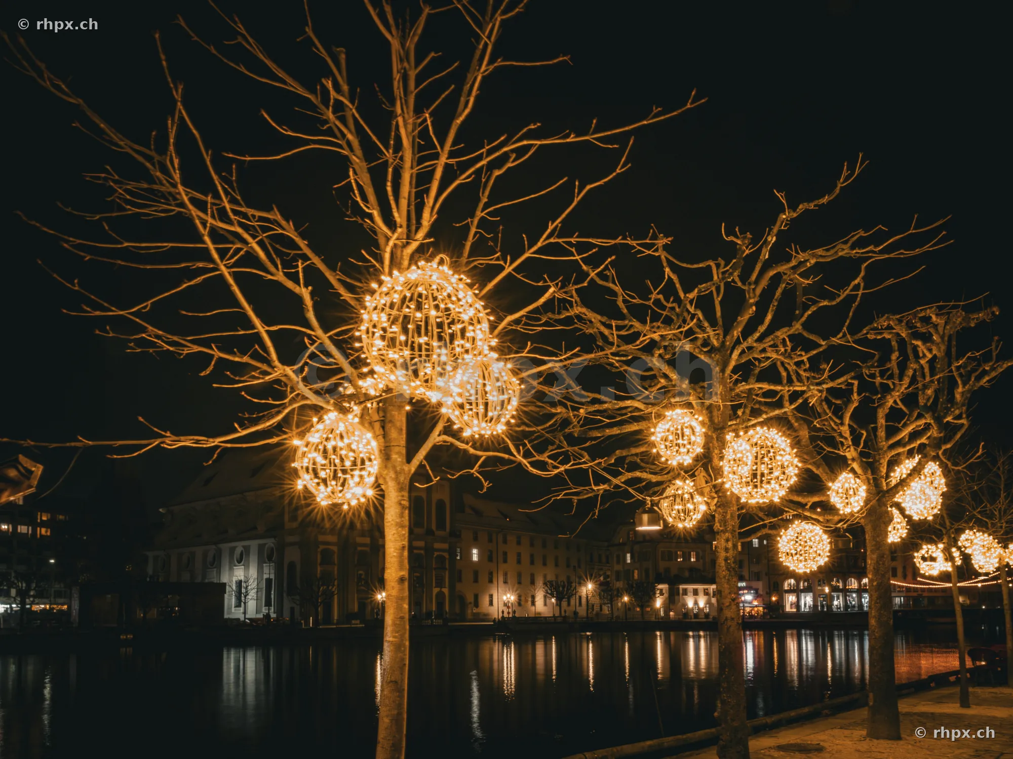 Christmas decorations with glowing spheres in the trees along the Reuss river in Lucerne at night.