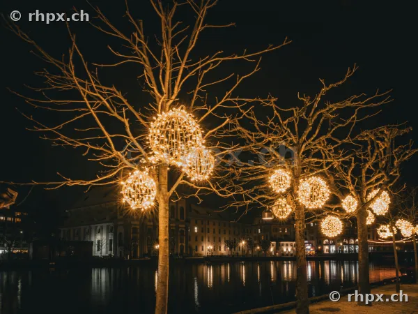Christmas decorations with glowing spheres in the trees along the Reuss river in Lucerne at night.