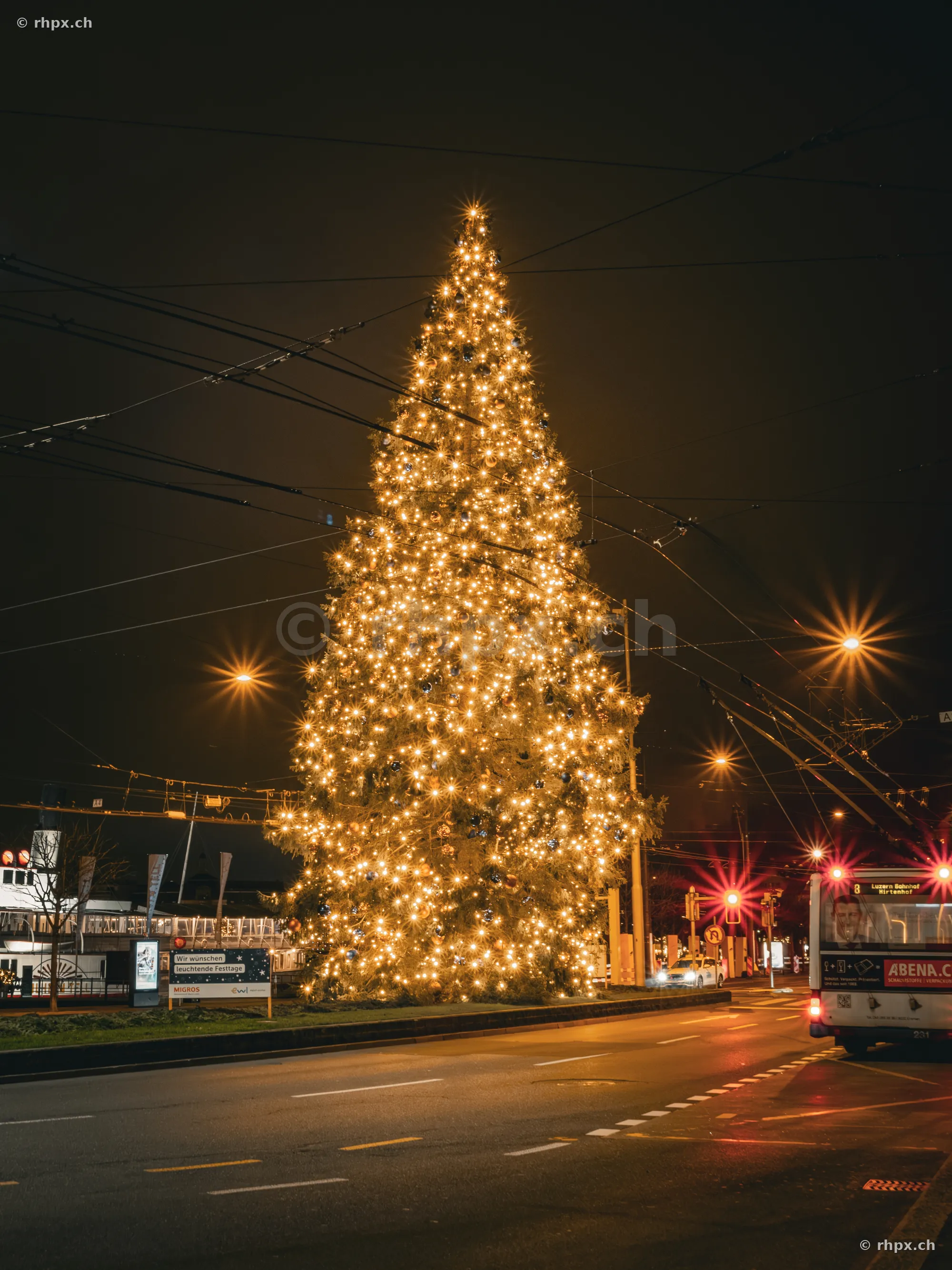 Illuminated Christmas tree in front of Lucerne train station at night with a bus passing by