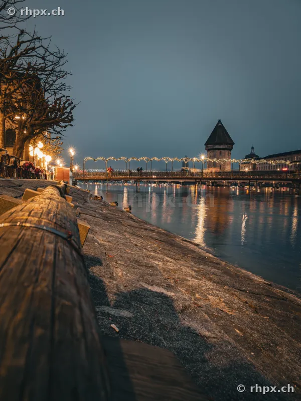 View along the Reuss river in Lucerne with Christmas lights on the bridge and the water tower at twilight