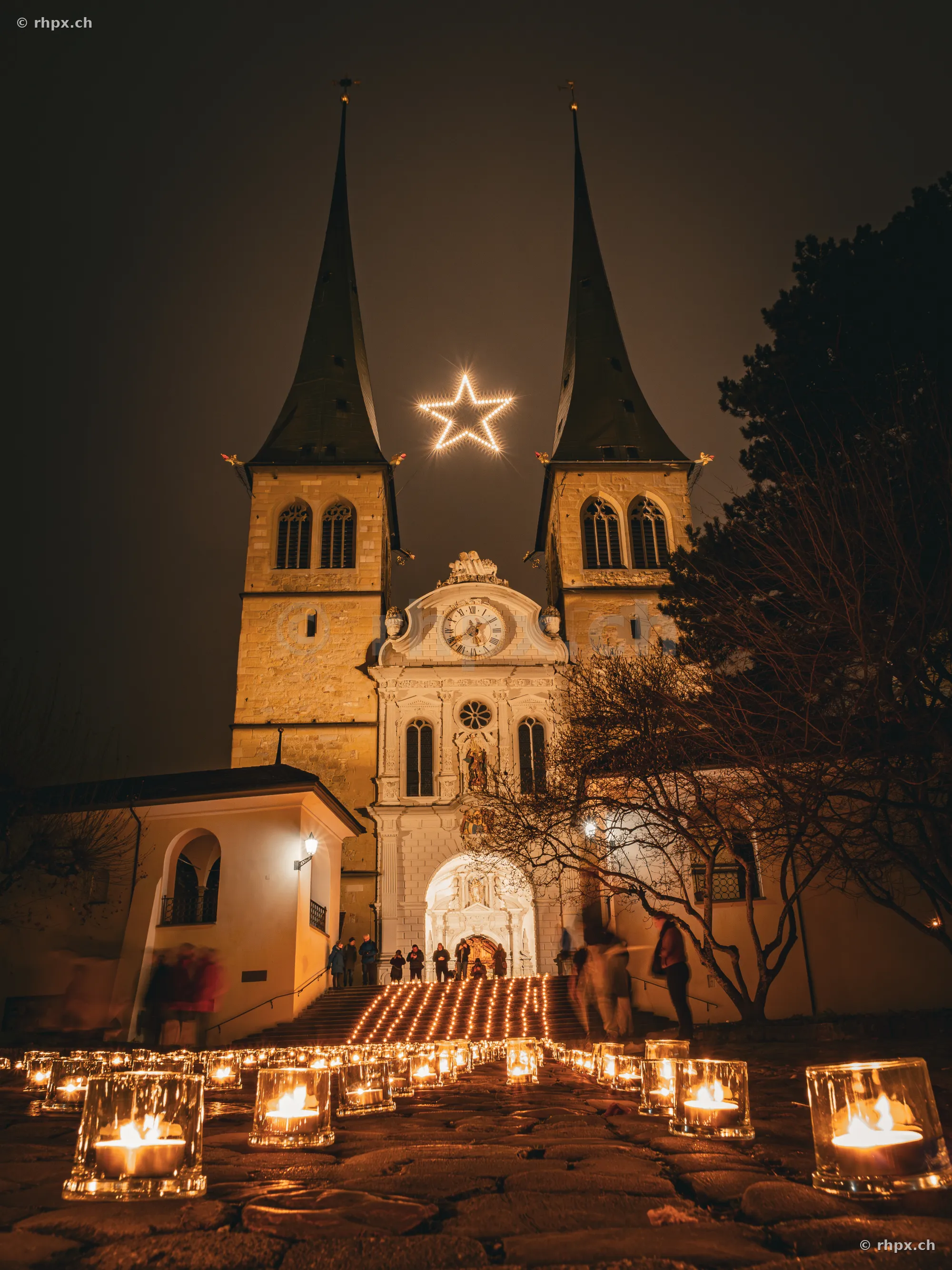 The illuminated Hofkirche in Lucerne with a glowing Christmas star between the towers and candles on the forecourt