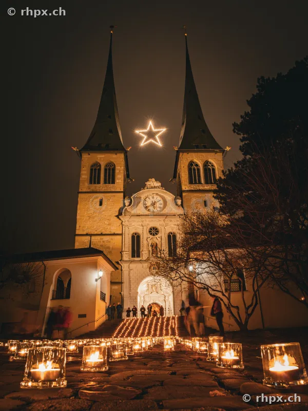 The illuminated Hofkirche in Lucerne with a glowing Christmas star between the towers and candles on the forecourt