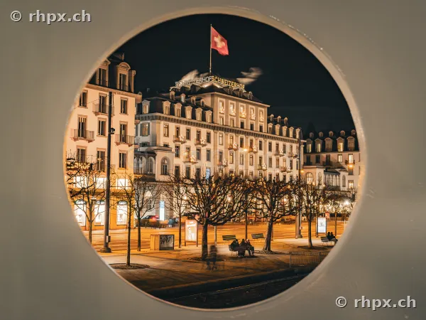 Blick durch ein rundes Fenster auf das beleuchtete Hotel Schweizerhof in Luzern.