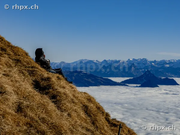 A person sits on the grassy slope of Mount Pilatus looking over a sea of clouds towards the Alps