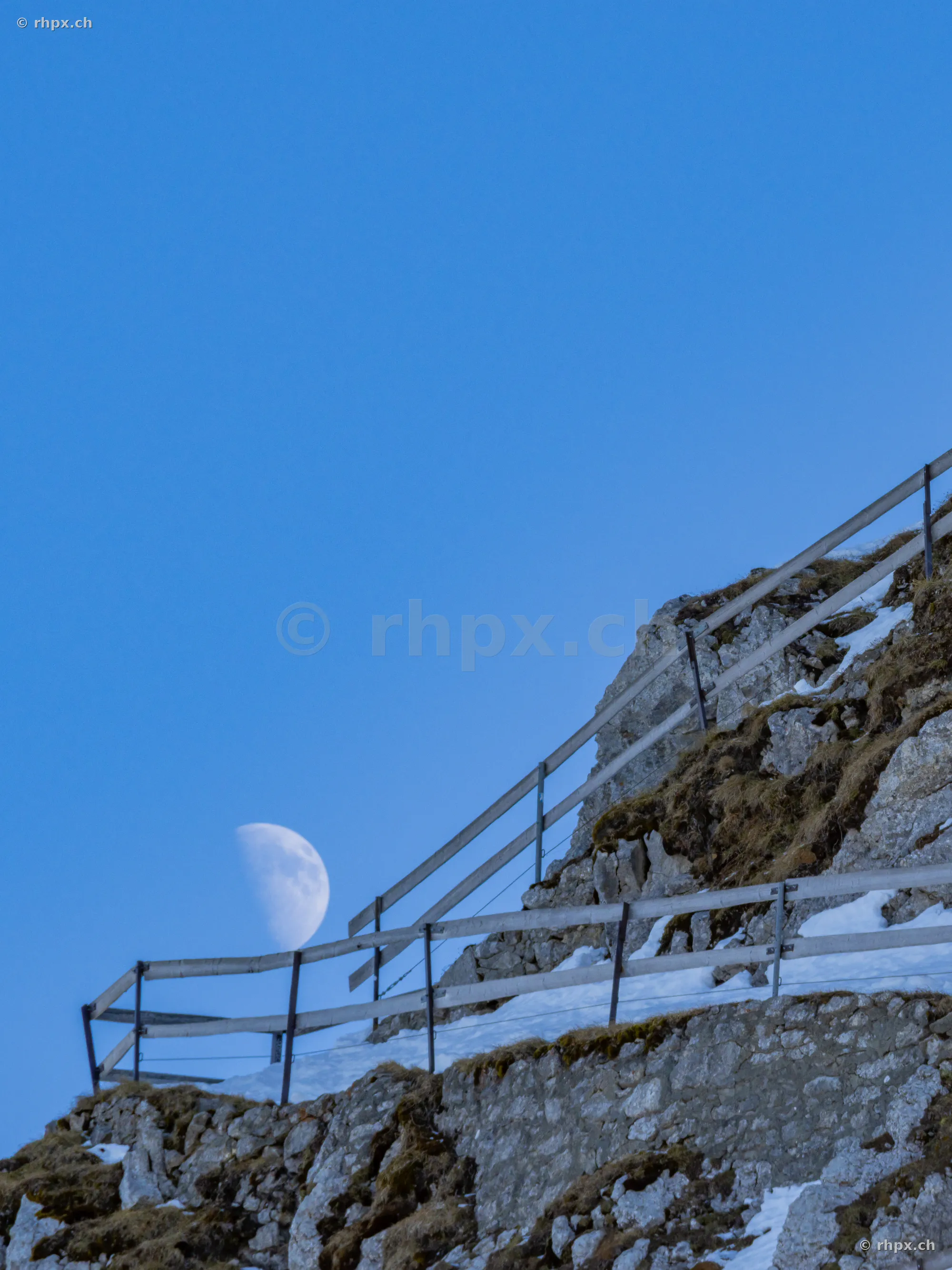 Rocky ridge of the Pilatus with a wooden fence and visible half moon in the blue sky