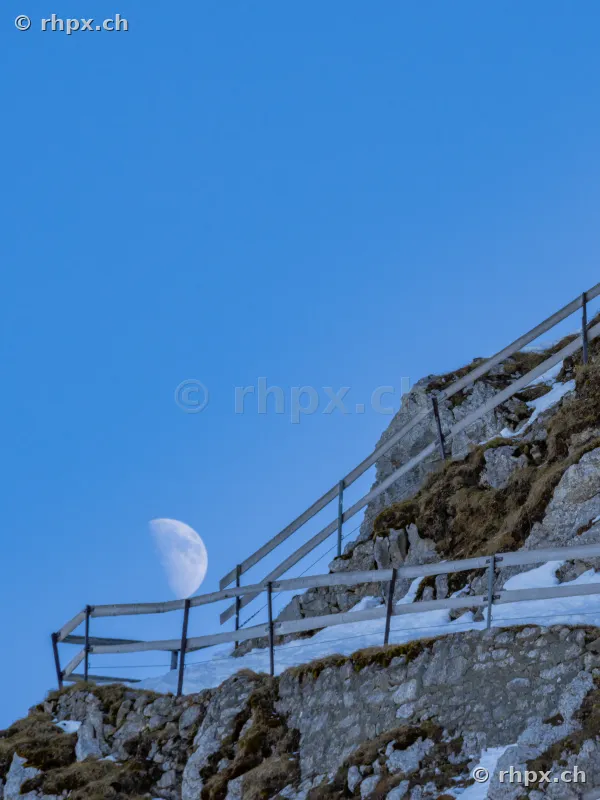 Rocky ridge of the Pilatus with a wooden fence and visible half moon in the blue sky