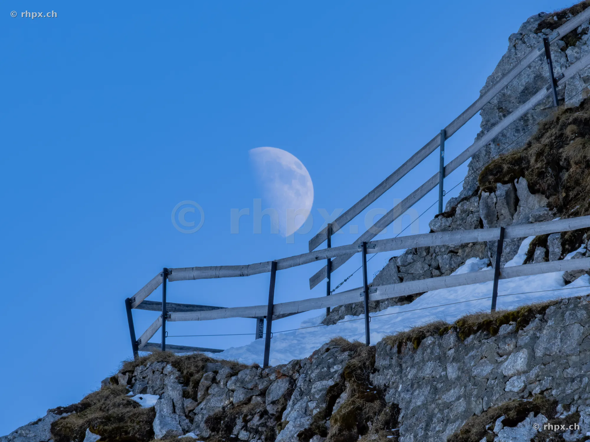 The waxing half moon in the blue daytime sky over a rocky path on Mount Pilatus with wooden fence and snow