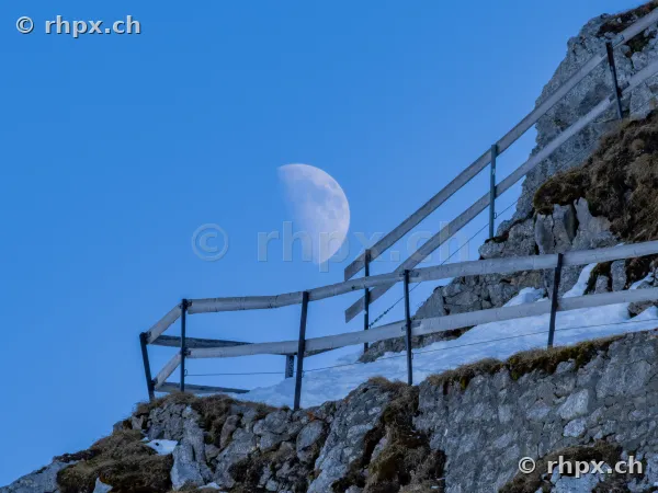 The waxing half moon in the blue daytime sky over a rocky path on Mount Pilatus with wooden fence and snow