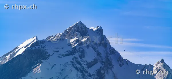 Felsiger, schneebedeckter Gipfel des Pilatus mit einer Seilbahnkabine vor blauem Himmel.