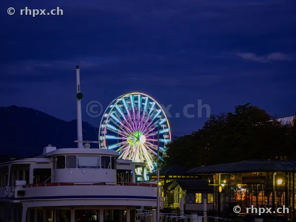 Illuminated Ferris wheel of the Lucerne Autumn Fair at night in front of a passenger ship