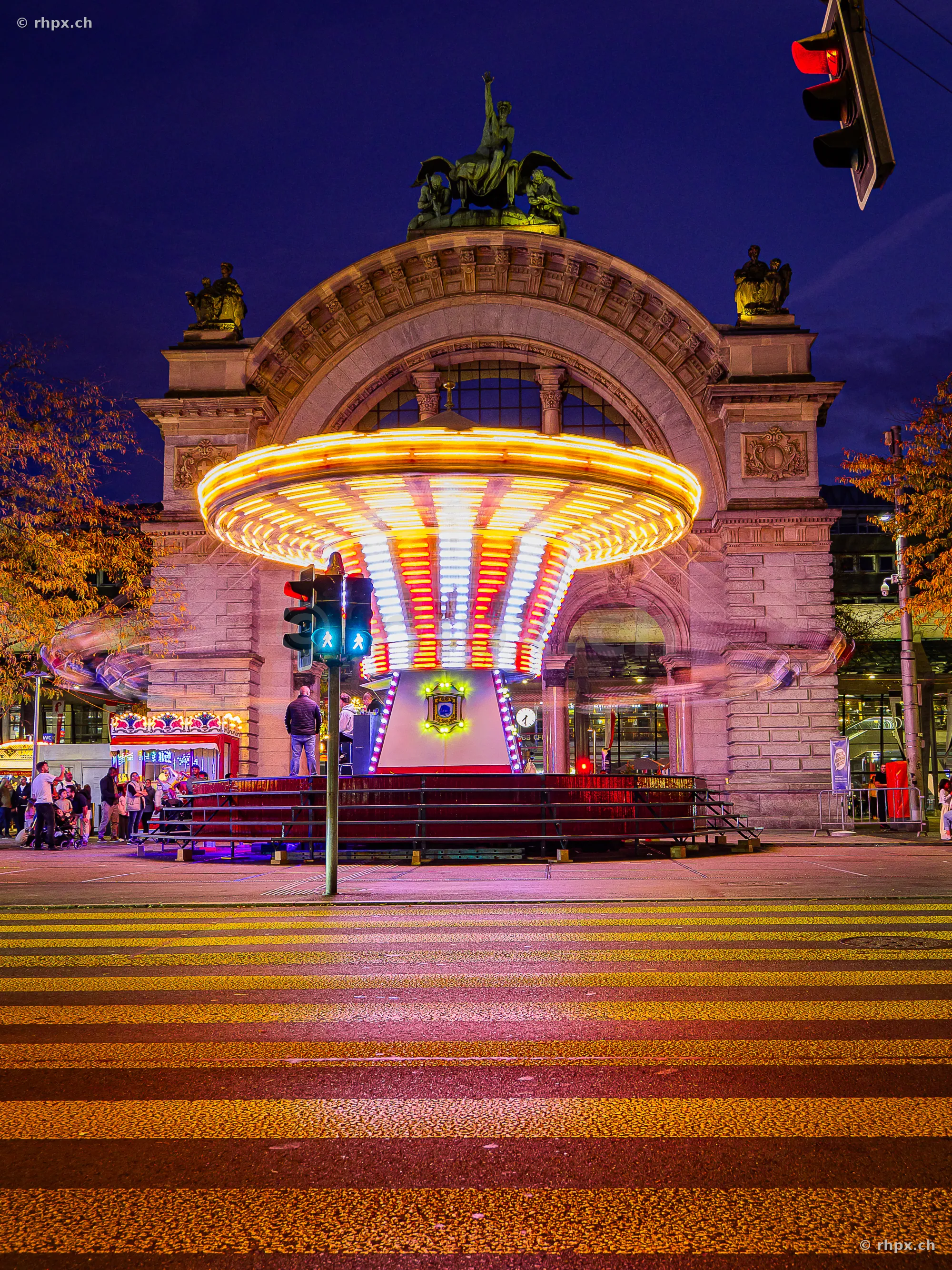Ein beleuchtetes Kettenkarussell der Lozärner Määs vor dem historischen Bahnhofsportal in Luzern bei Nacht.
