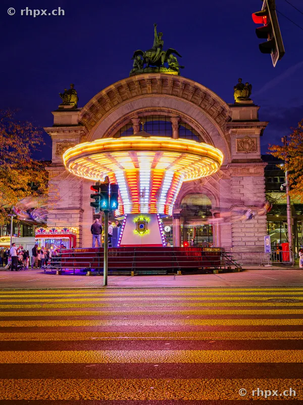 An illuminated chain carousel at the Lozärner Määs in front of the historic train station portal in Lucerne at night.