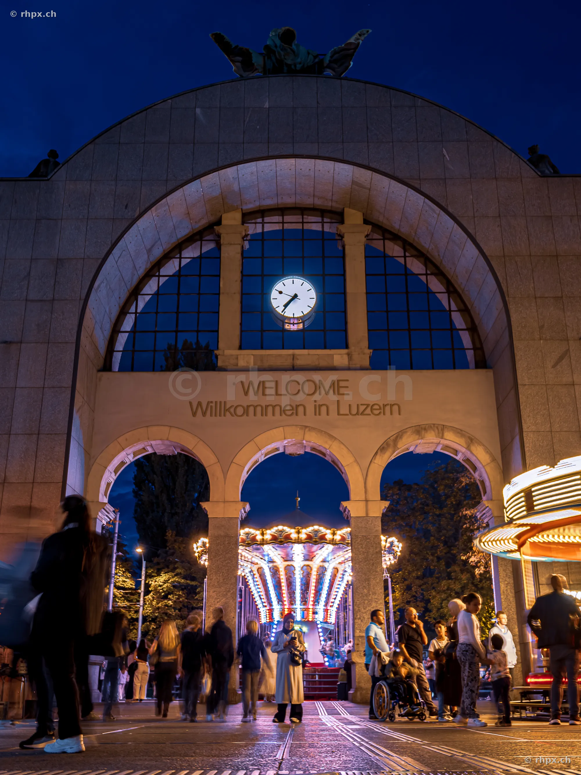 Lucerne station gate in the evening with a view of an illuminated fairground carousel.