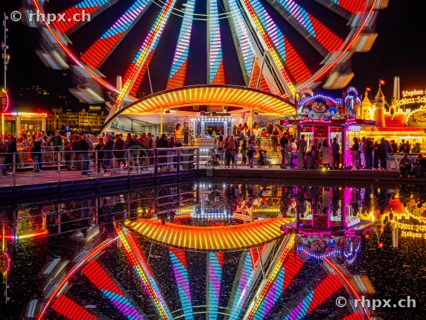 Ferris wheel and fairground stalls at the Lucerne autumn fair at night with reflection in water