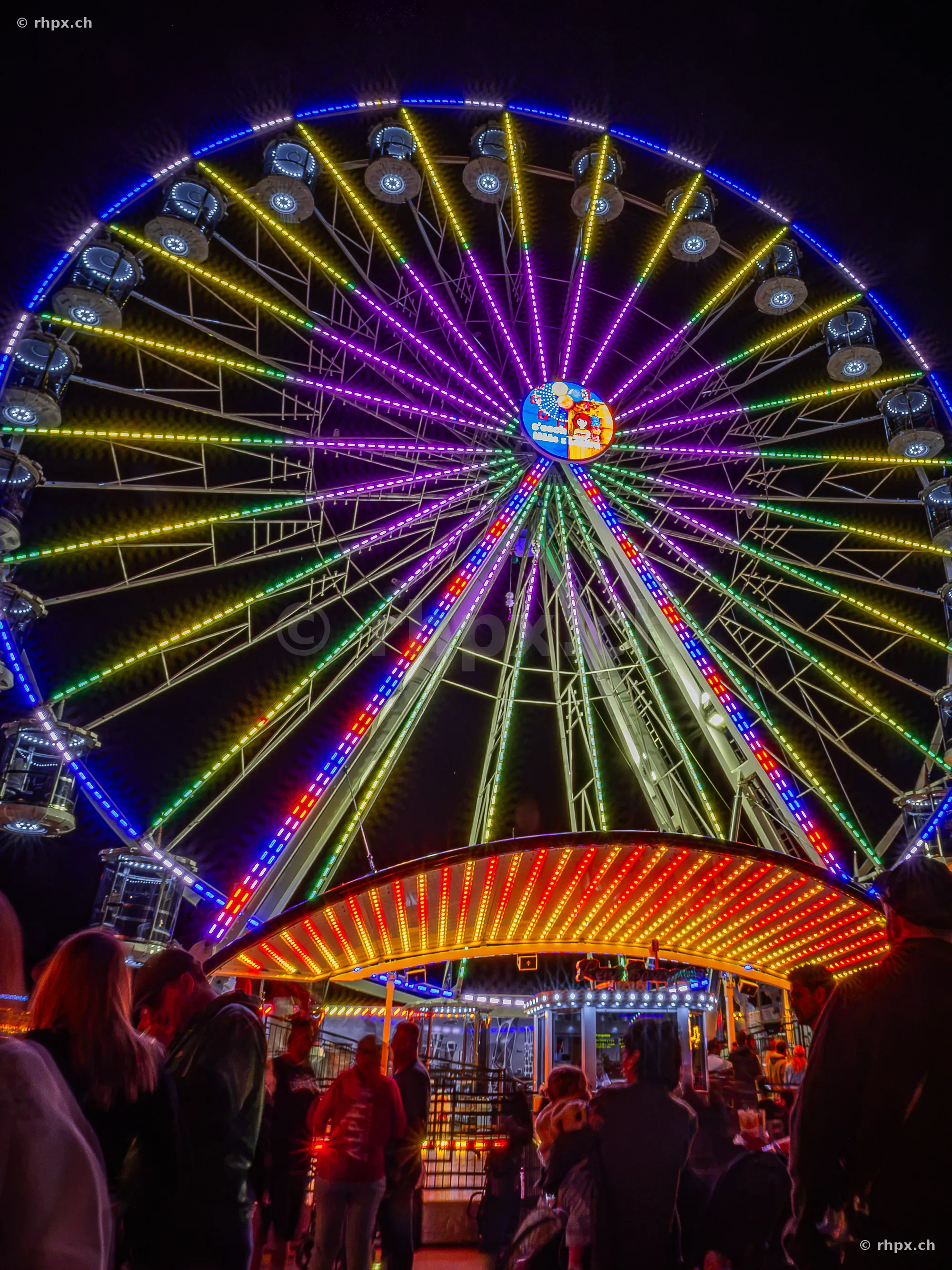 Illuminated Ferris wheel at the Lozärner Määs at night with people in the foreground.