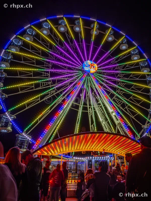 Illuminated Ferris wheel at the Lozärner Määs at night with people in the foreground.